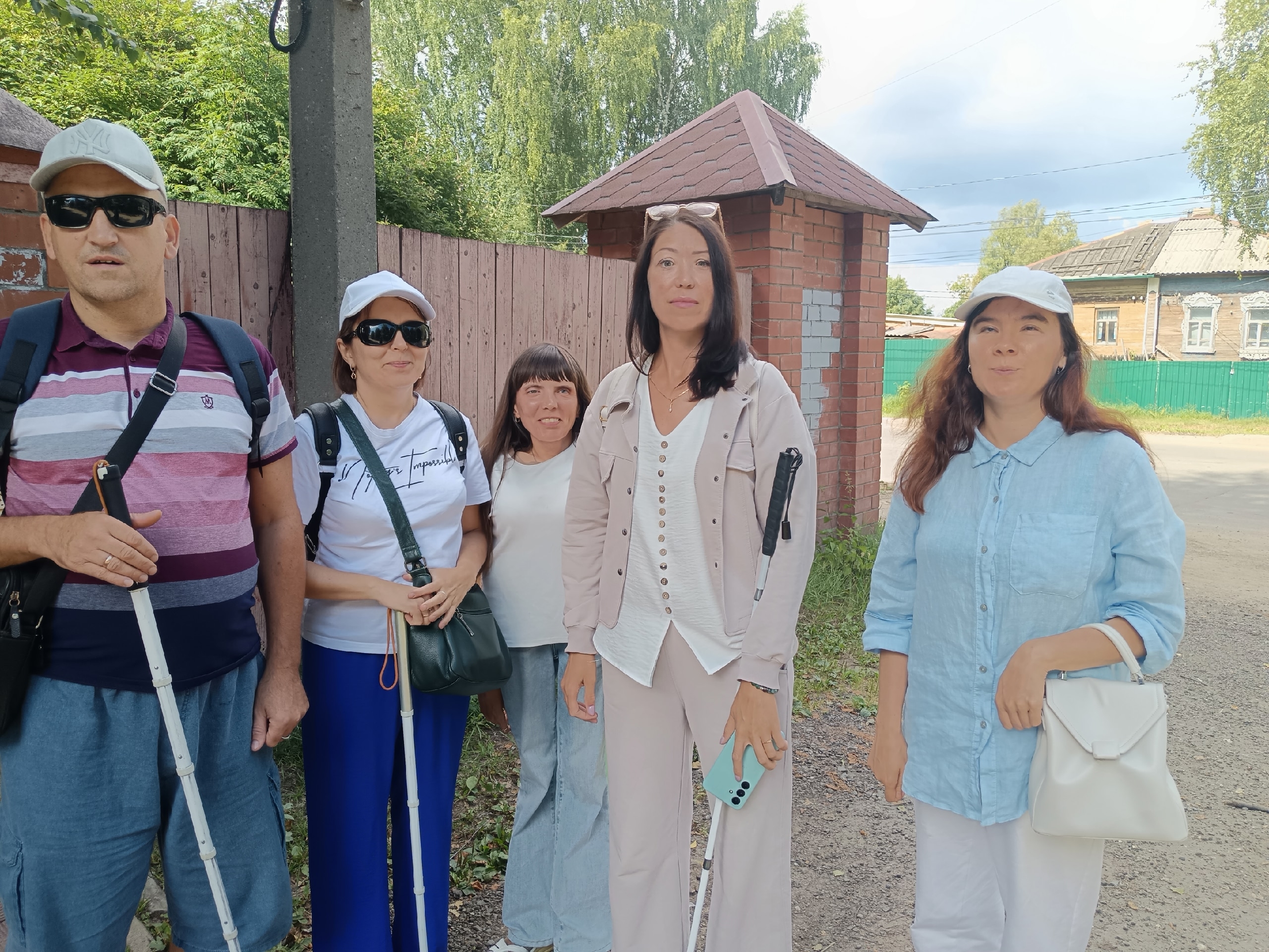 Группа людей с тактильными тростями для незрячих стоит на улице около деревянного забора