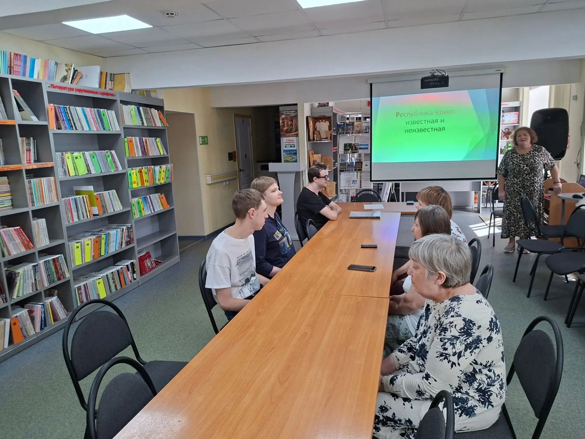 Лектор выступает перед слушателями в библиотеке на фоне экрана презентации