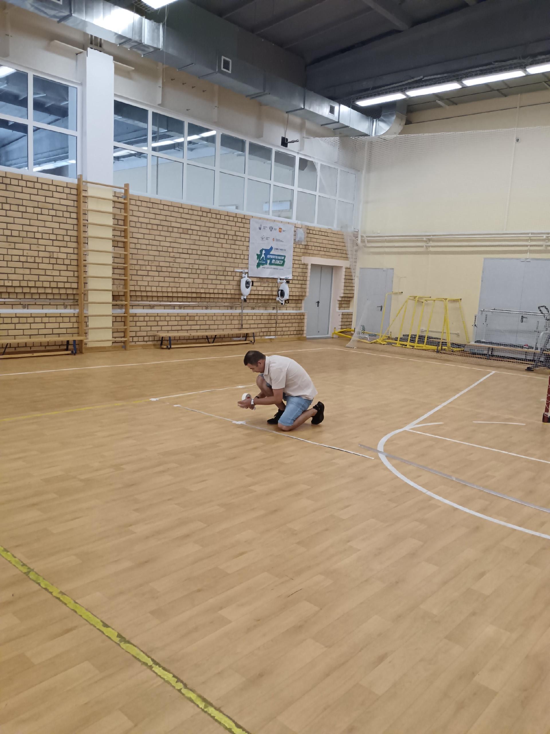 Мужчина на коленях наклеивает белую ленту для разметки площадки на деревянный пол