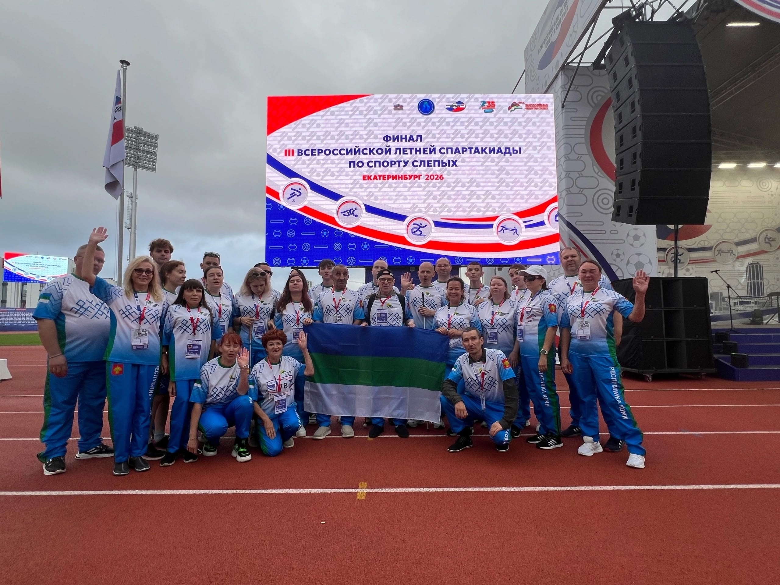 Группа спортсменов в сине-белой форме позирует с флагом на фоне главного экрана спартакиады
