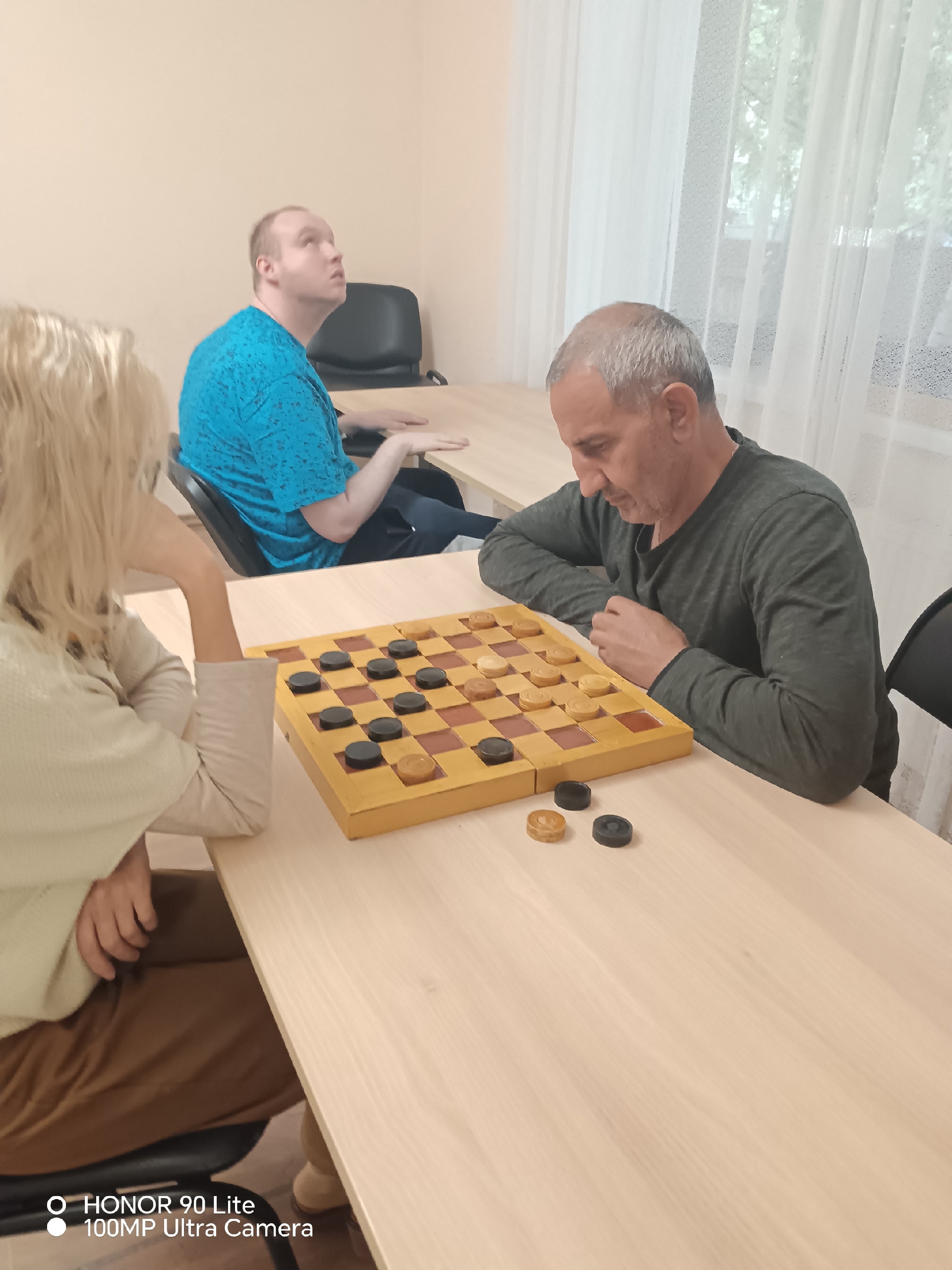 Мужчина и светловолосая женщина играют в шашки за длинным деревянным столом
