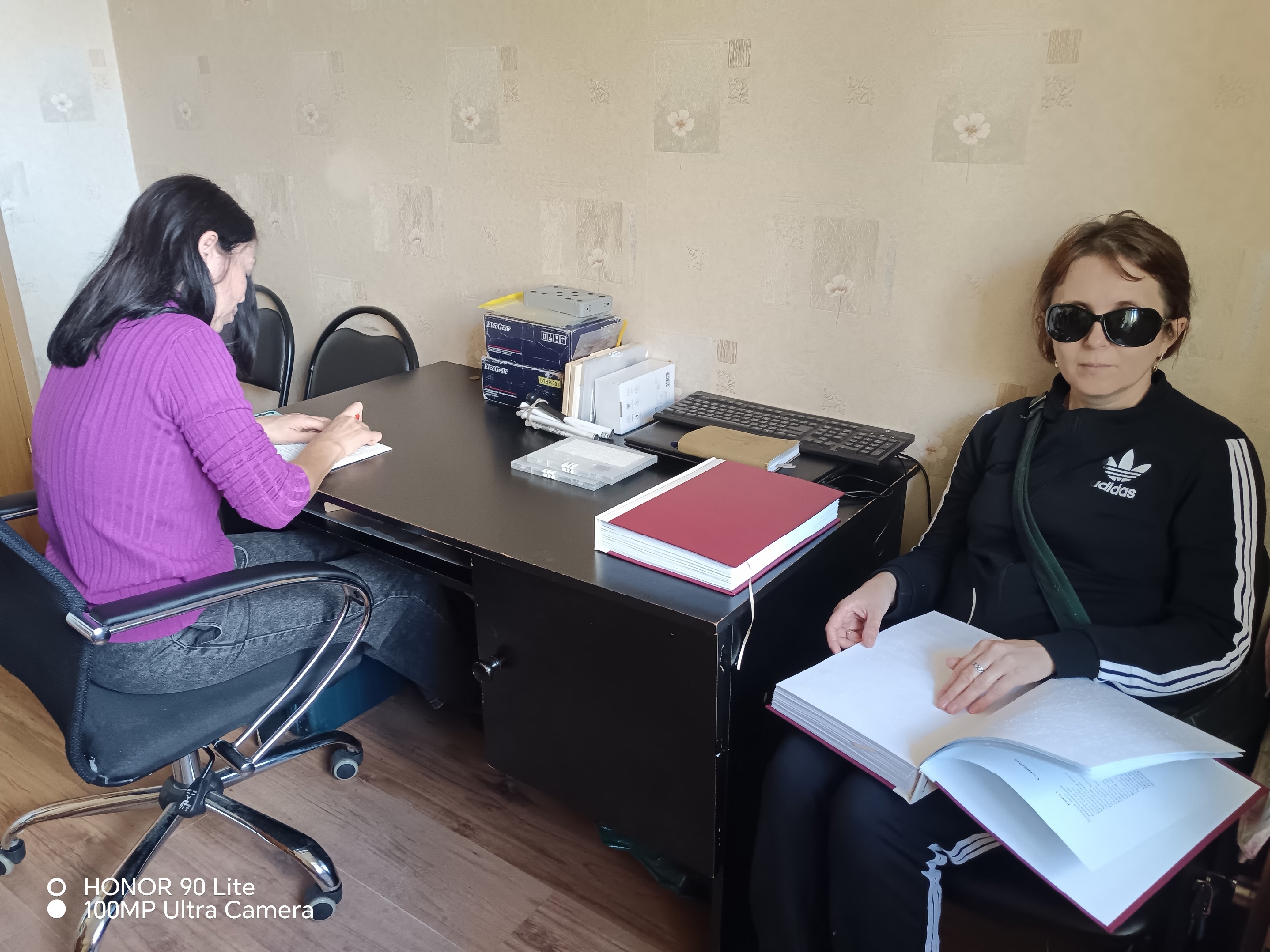 Женщина в темных очках с открытой книгой по Брайлю сидит у рабочего стола