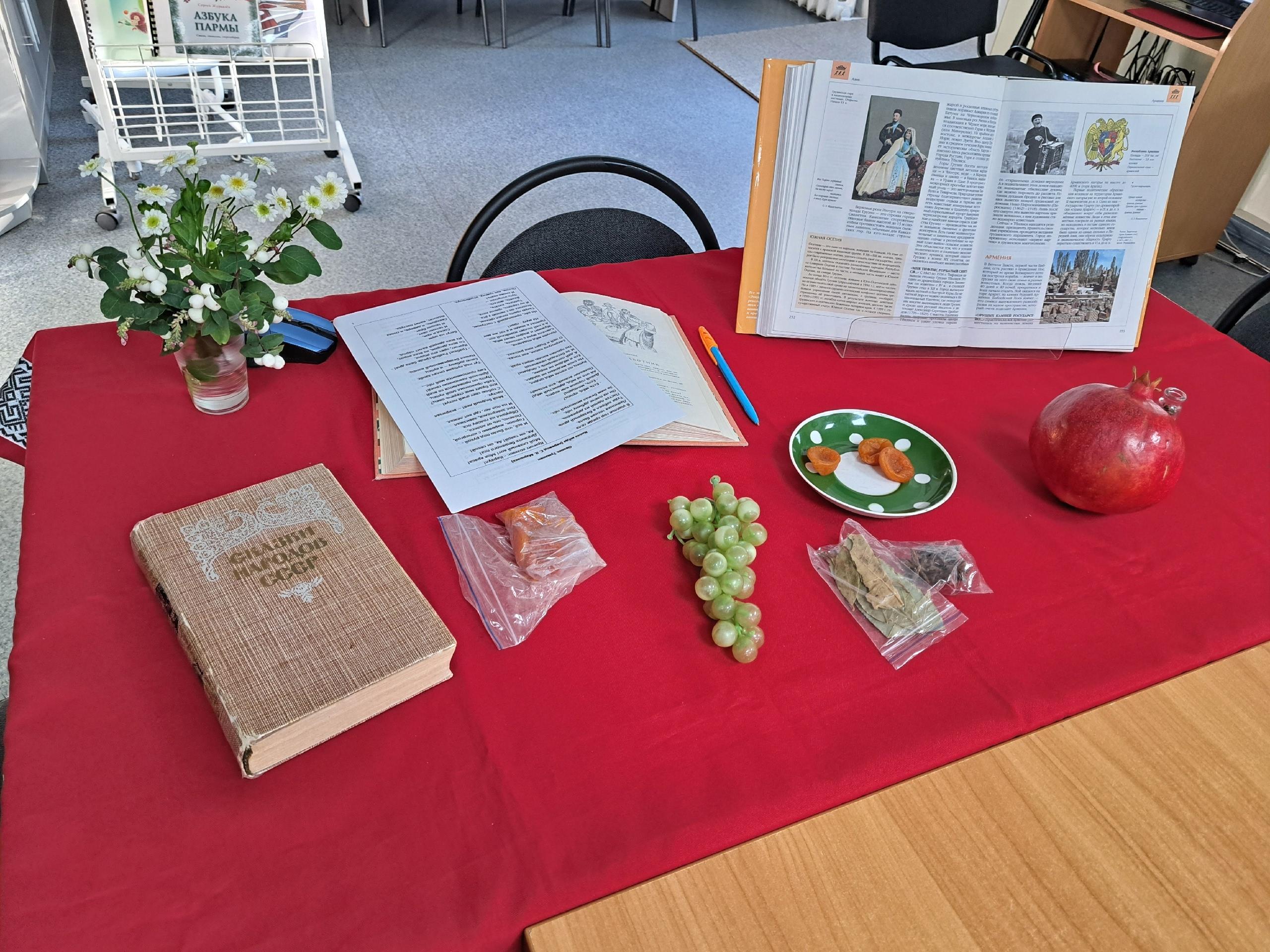 Стол, накрытый красной скатертью, с книгами, открытой книгой, гранатом, виноградом и сухофруктами