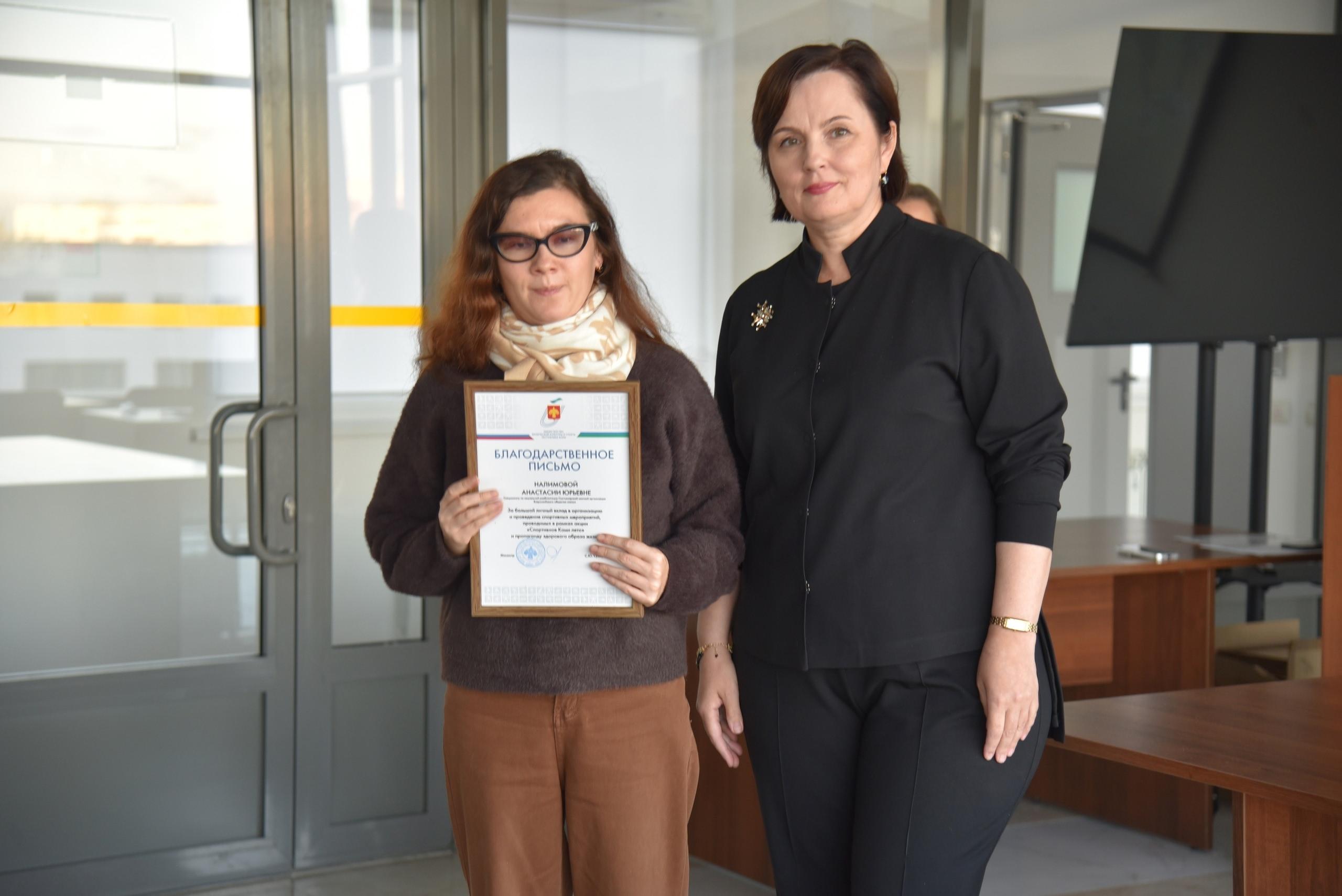 Министр спорта вручает благодарственное письмо женщине в очках и свитере