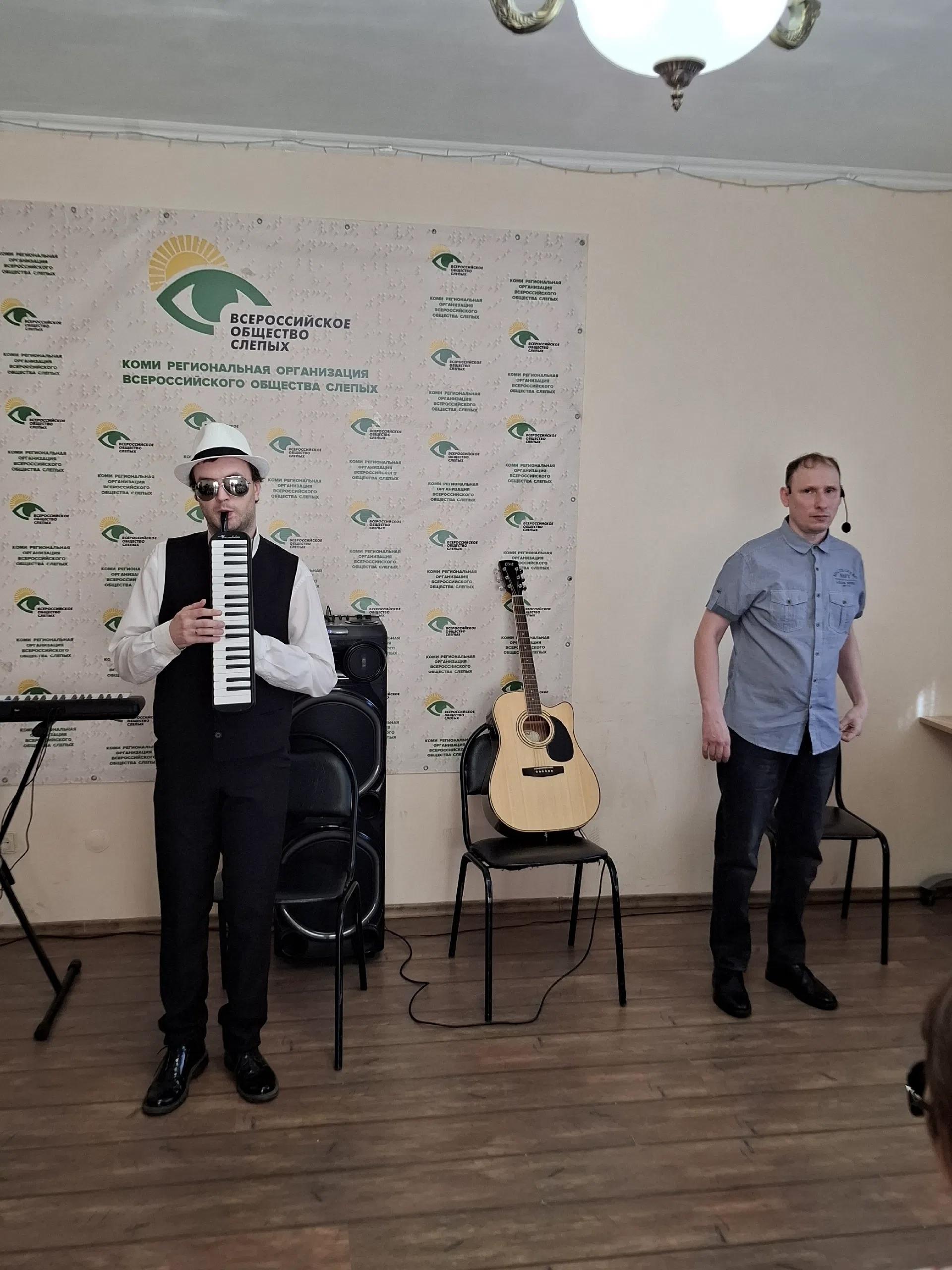 Музыкант в шляпе и очках играет на мелодике на фоне фирменного баннера организации